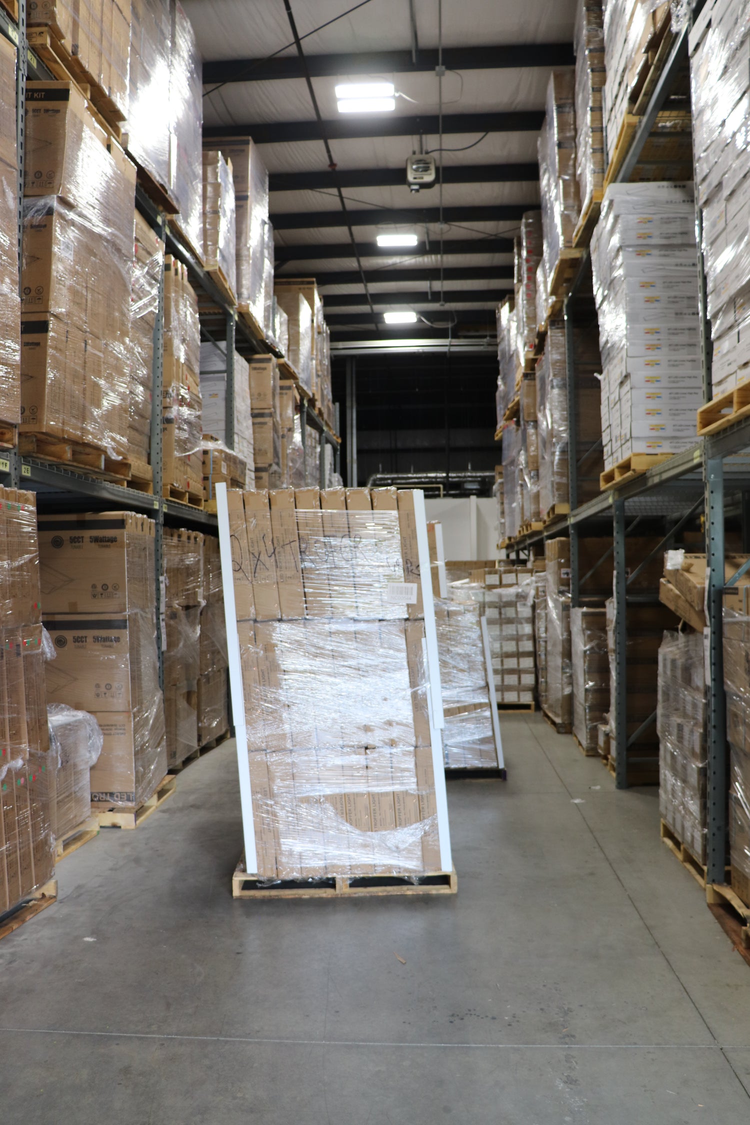Warehouse aisle with NCLTG LED high bay lights illuminating stacked pallets of products ready for shipment